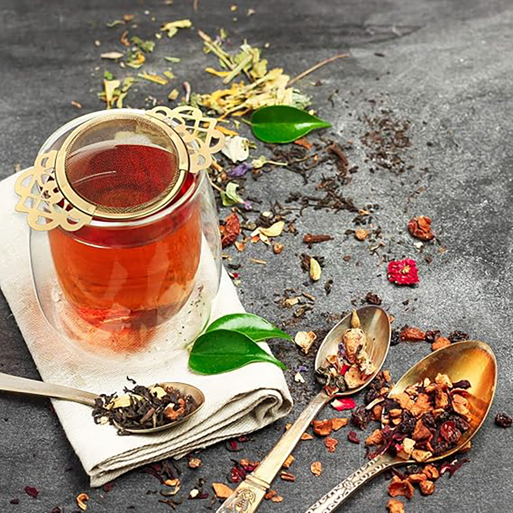 A gold tea strainer sits in a clear glass with tea steeped inside. Spoons with loose leaf tea on the are on the table with a cloth napkin