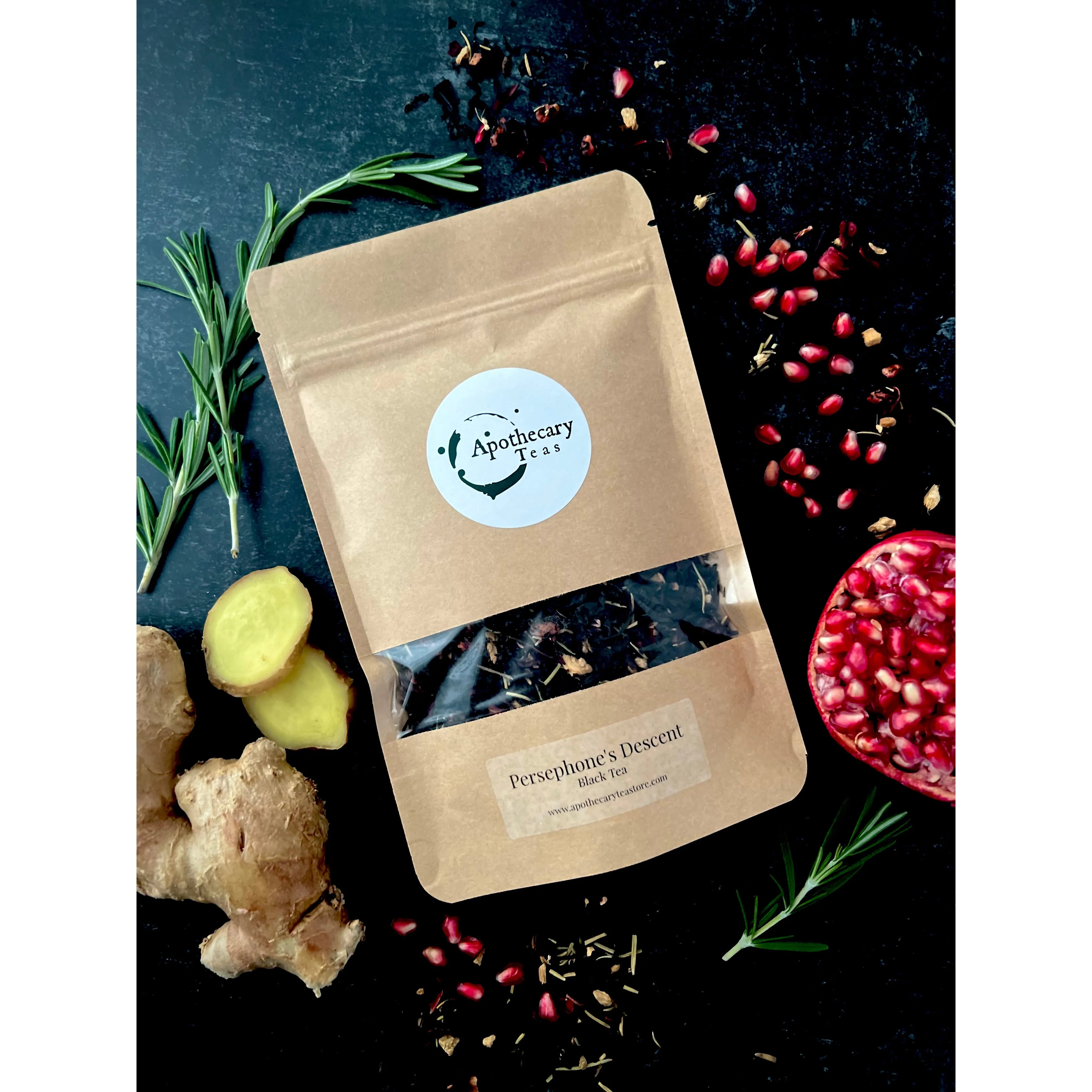 A brown bag of Persephone's Descent black tea surrounded by fresh ginger and rosemary with half of a pomegranate and pomegranate arils spread around it